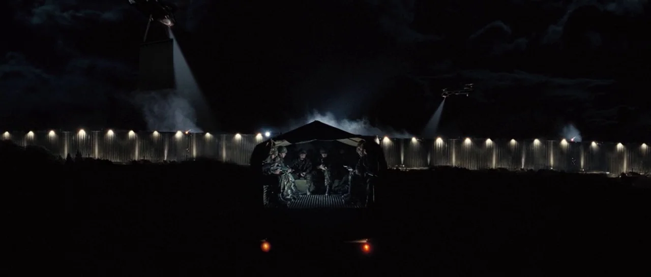 Doomsday (2008) movie still: wide — A dark night landscape featuring a long illuminated perimeter wall in the distance. Soldiers sit in the back of…
