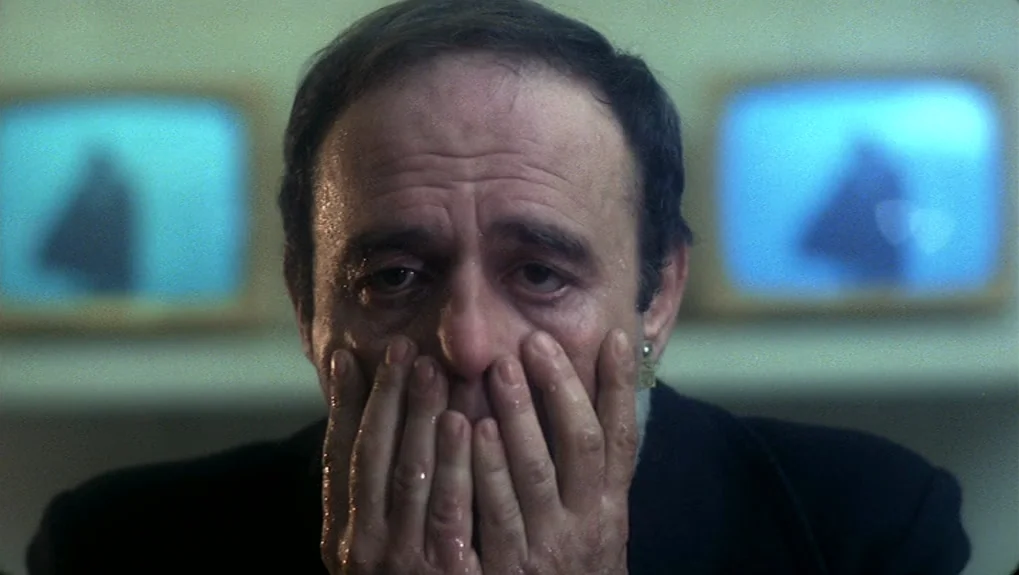The War of the Worlds: Next Century (1981) movie still: closeup — A man stands before two glowing television screens that cast a cool blue light. He…