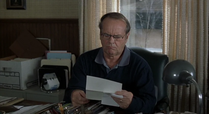 About Schmidt movie still: medium — A cluttered personal workspace features a paper-filled desk and a box of files. An older man sits in a black…