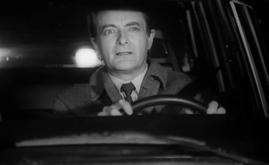Vivement Dimanche (1983) movie still: medium — A man drives a car at night in a dimly lit vehicle cabin. Bright headlights from an unseen trailing…