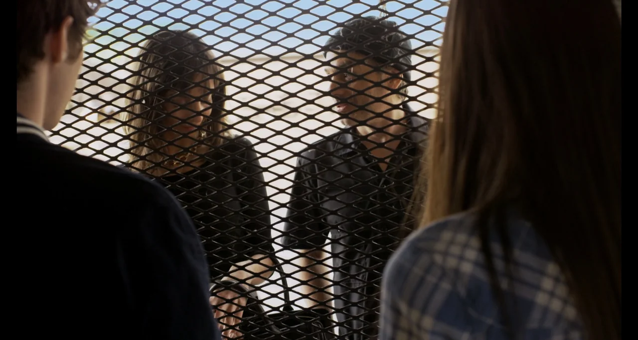 A.R.C.H.I.E. (2016) movie still: medium — A man and a woman stand positioned behind a diamond-patterned metal security gate. The two subjects appear…