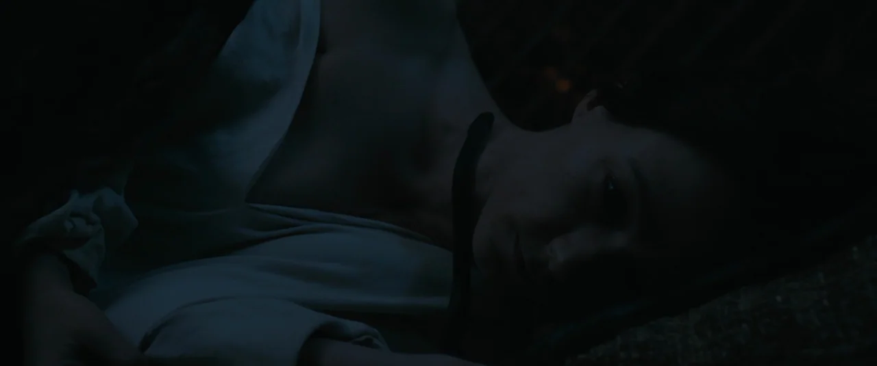 Hagazussa (2017) movie still: closeup — A dark, dimly lit bedroom environment with the subject resting on a textured surface. A woman in a white…