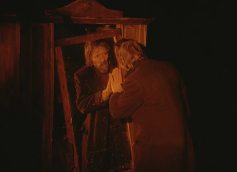 Russian Symphony (1994) movie still: medium — A man stands in a dark room before a weathered, freestanding wooden floor mirror. He presses his palm…