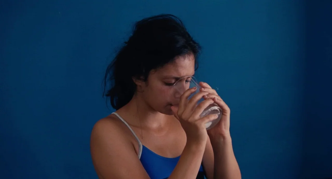 Ava (2017) movie still: medium — A woman stands against a flat, solid dark blue wall indoors. She holds a clear glass with both hands and drinks…