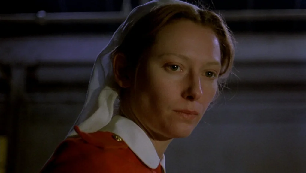 War Requiem (1989) movie still: closeup — A dimly lit hospital ward interior features a woman in a vintage uniform. The woman stands against a dark,…