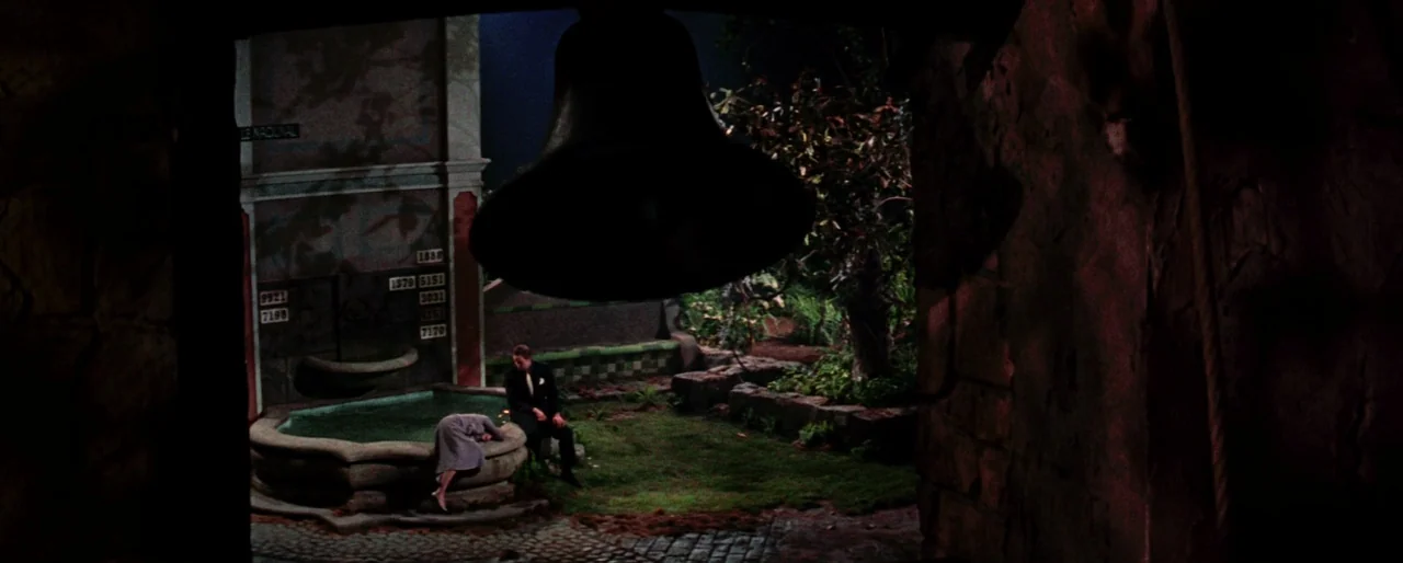 Guys and Dolls (1955) movie still: wide — A night time stone courtyard features a central water basin and stone walls. A man sits by the water while…