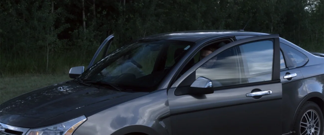 Radius (2017) movie still: medium — A grey sedan sits parked on the edge of a road bordered by woods. A man sits inside the vehicle with the…