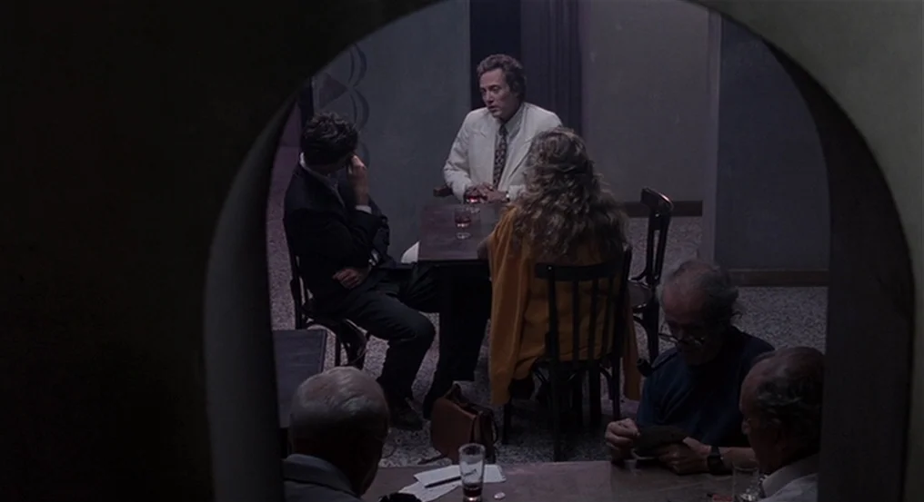 The Comfort of Strangers (1990) movie still: wide — A dimly lit room with stone-like walls seen through a dark arched doorway. Five people occupy…