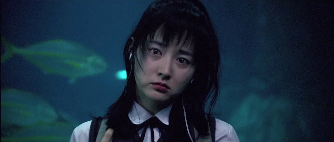 Lady Vengeance (2005) movie still: closeup — A young woman stands in front of a dark blue aquarium tank filled with fish. She wears wired earphones…