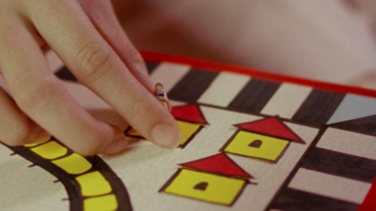 Shirkers (2018) movie still: closeup — A close-up shows a human hand manipulating a tiny figurine on a colorful board. The game board features paper…
