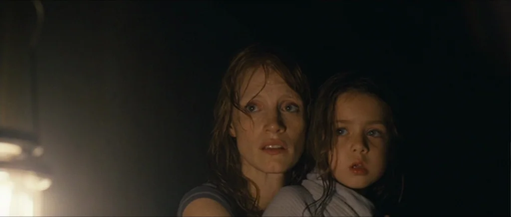 Take Shelter (2011) movie still: medium — A woman and a young child stand together in a dark, shadow-filled room. They look toward the left side of…