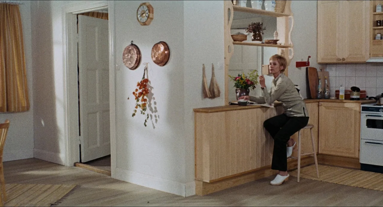 The Touch (1971) movie still: wide — A bright kitchen features light wood cabinets and decorative wall hangings on white walls. A woman sits on a…