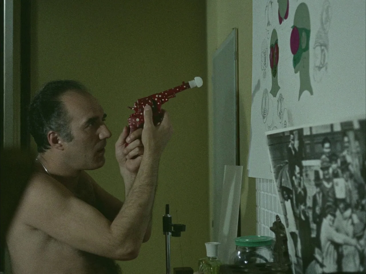 Dillinger is Dead (1969) movie still: medium — A dimly lit room features walls covered with schematic sketches and black-and-white photographs. An…