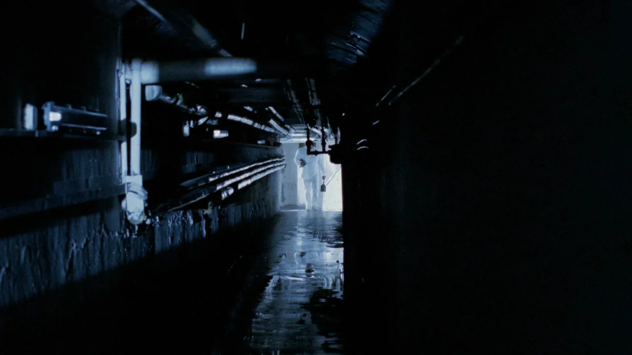 The Sect (1991) movie still: wide — A dark subterranean utility tunnel contains overhead pipes and reflective puddles on the ground. A single person…