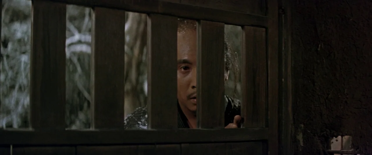 Ballad Of Narayama (1958) movie still: closeup — A dark wooden gate with vertical slats obscures part of a man's face. He looks through the gaps…