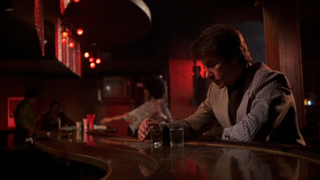 Mean Streets (1973) movie still: medium — A dimly lit 1970s lounge features a polished wooden counter and red hanging lights. A man sits at the bar…
