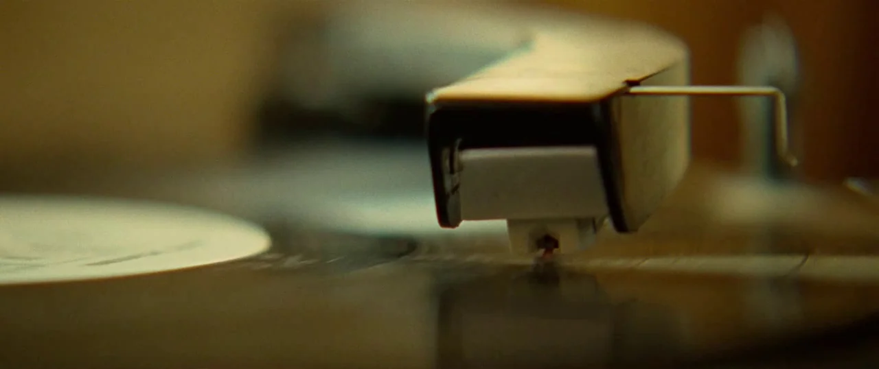 Low Down (2014) movie still: closeup — A close-up view of a vinyl record player needle resting on a spinning disc. The cartridge and part of the…
