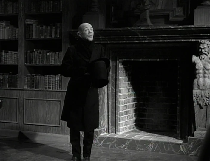 The Devil’s Eye (1960) movie still: fullbody — A dimly lit room features floor-to-ceiling bookshelves and a large stone fireplace. A bald man…