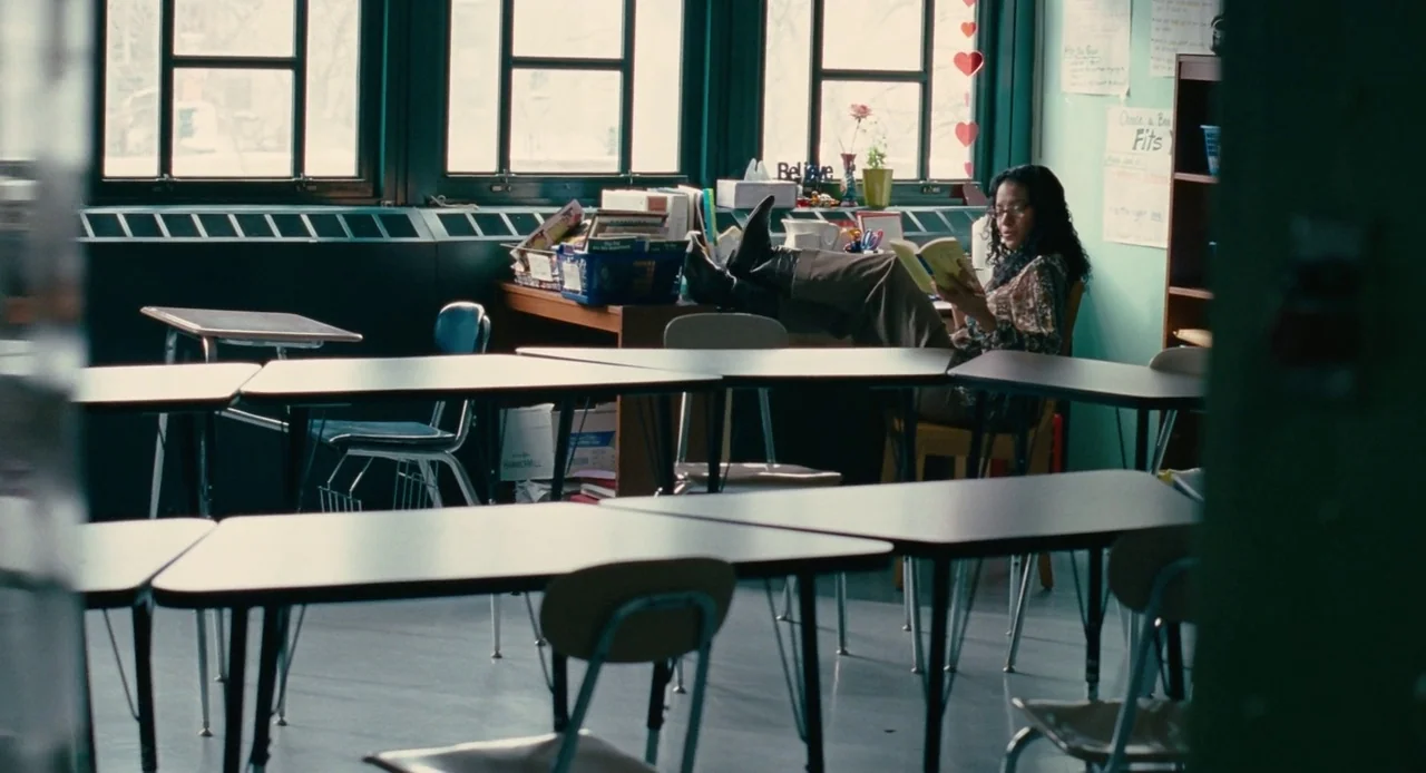 Pariah (2011) movie still: wide — An empty classroom contains rows of desks and chairs facing towards large windows. A teacher sits at her desk with…