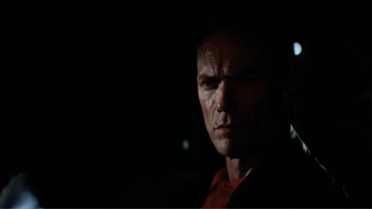 Heartbreak Ridge (1986) movie still: closeup — A dark vehicle interior with the man illuminated against a pitch black background. The man sits in…