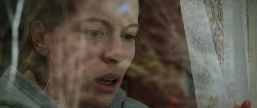 Snow Angels (2007) movie still: closeup — A woman stares through a glass pane with thin lace curtains hanging beside her. Reflection lines are…
