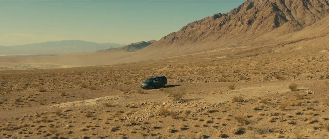 American Sicario (2021) movie still: wide — An arid desert landscape features sparse vegetation and rolling mountains under a clear sky. A lone SUV…