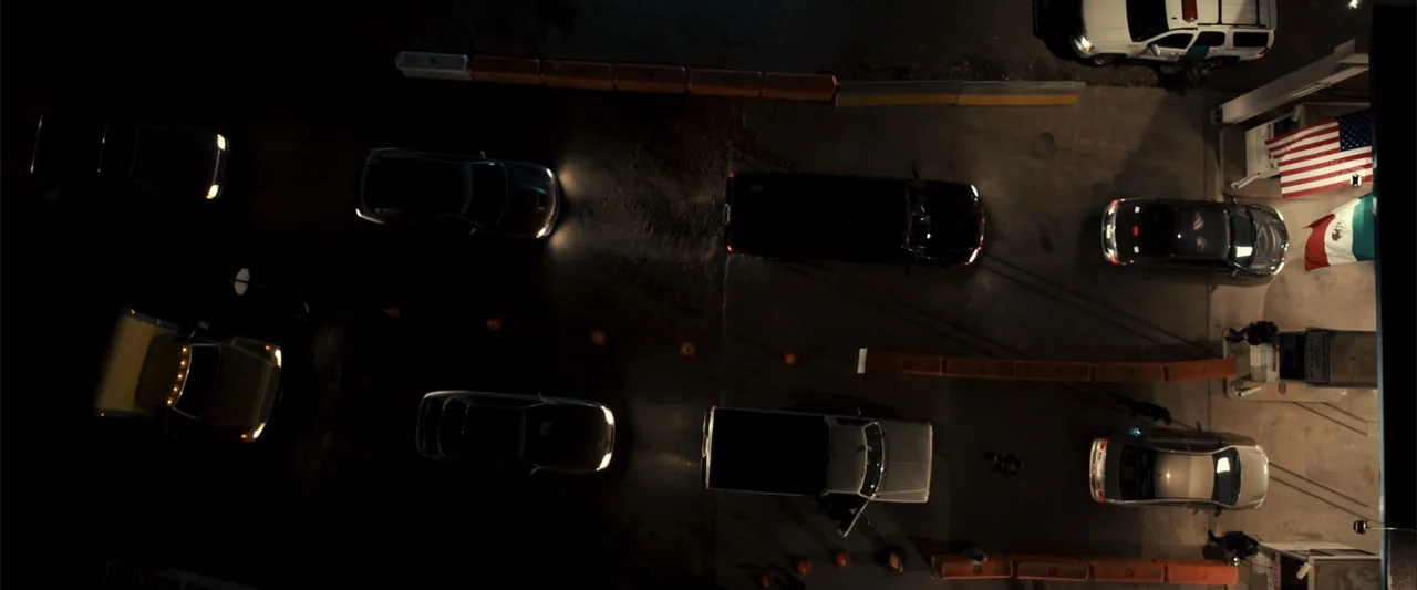 Frontera (2014) movie still: establishing — A dark outdoor border checkpoint at night features multiple lanes of stationary cars. Vehicles idle in…