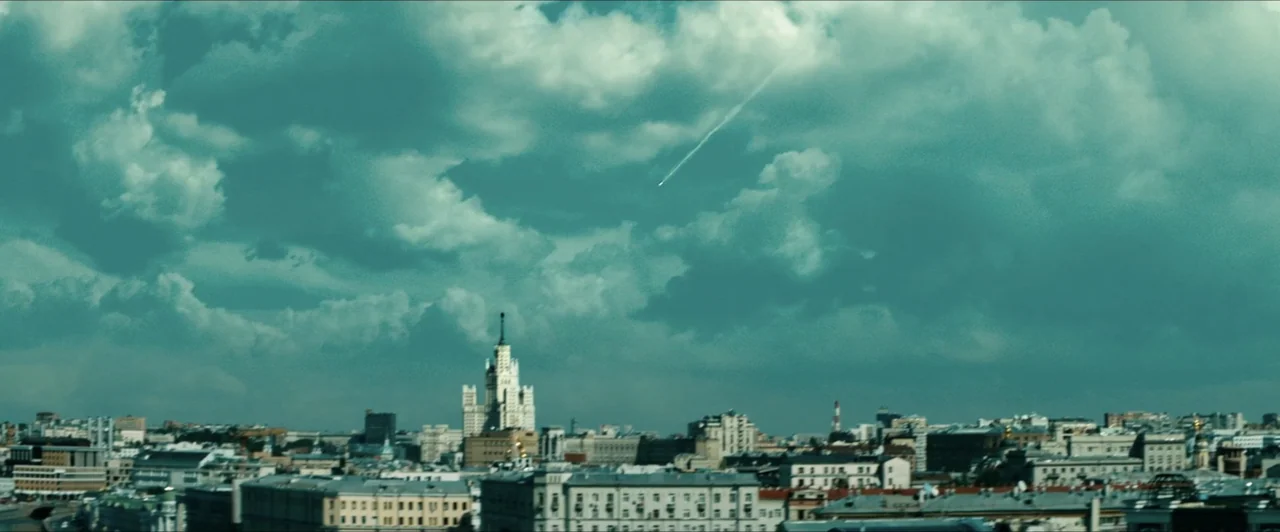 Invasion (2020) movie still: establishing — An aerial view of a dense city skyline dominated by a tall Soviet-era skyscraper. A white vapor trail…