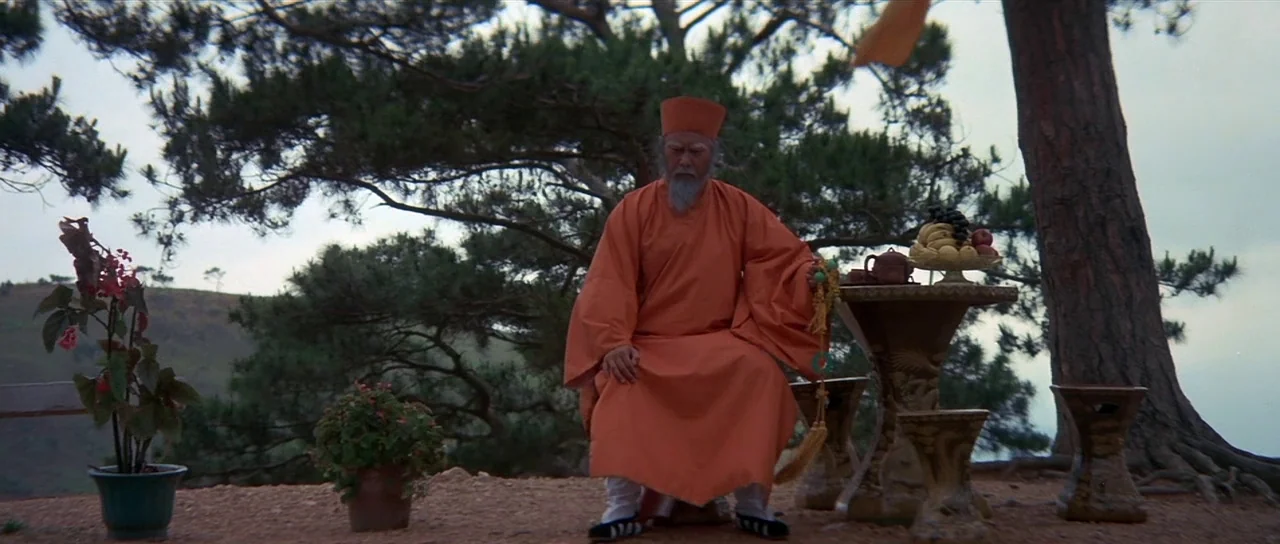 Enter The Dragon (1973) movie still: fullbody — A monk sits outdoors on a mountainous terrain beneath large, sprawling pine trees. He holds prayer…