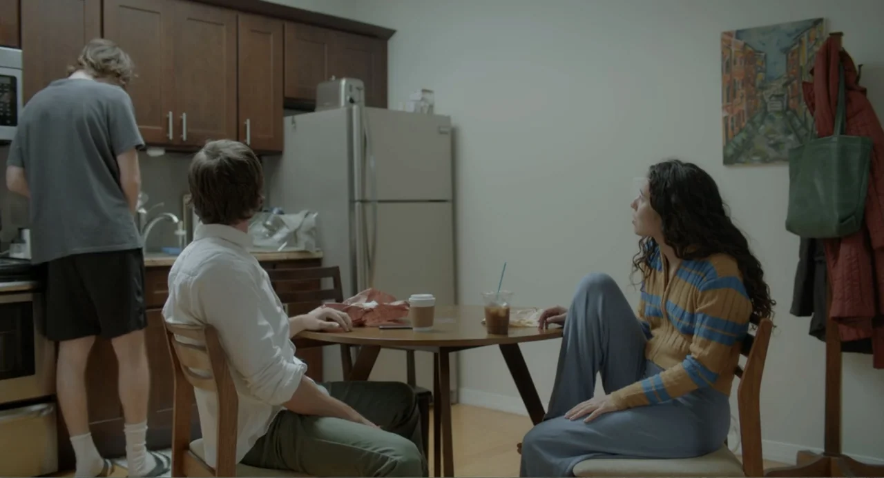This Closeness (2024) movie still: fullbody — A residential apartment with wood cabinetry and a round table occupied by three people. The subjects…