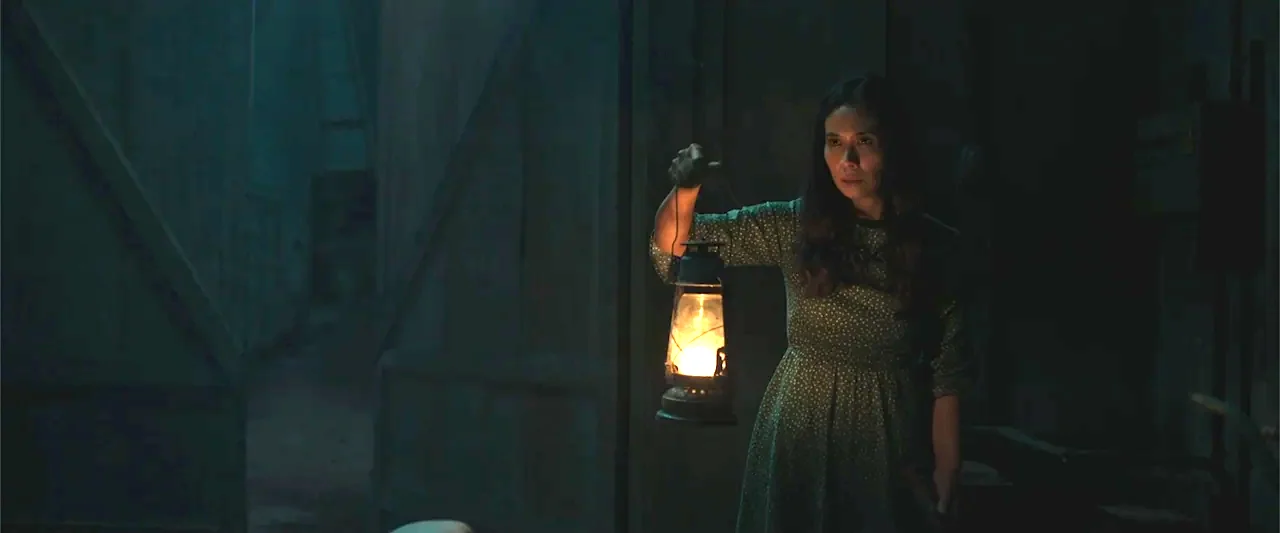 The Unholy (2022) movie still: medium — A dark, cluttered space with deep shadows and wooden structural elements visible in the background. A woman…