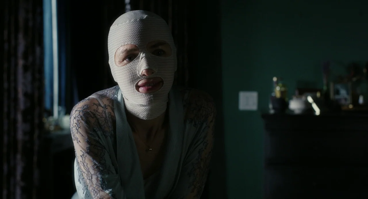 Goodnight Mommy (2022) movie still: medium — A dark, dimly lit room with sage green walls and blurred decorative objects in back. A woman with her…