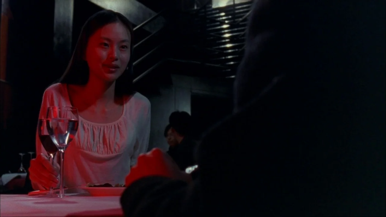 Audition (1999) movie still: medium — A dark restaurant interior with a table set for two people. A woman in a white shirt sits opposite an unseen…