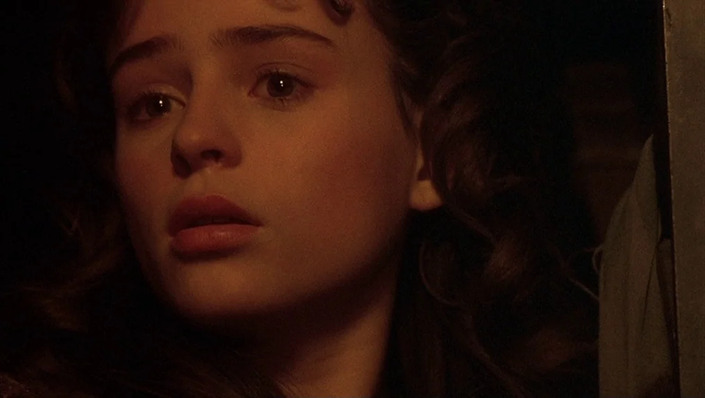 The Company of Wolves (1984) movie still: closeup — A dimly lit interior space with dark shadows concealing the surroundings. A young woman with…