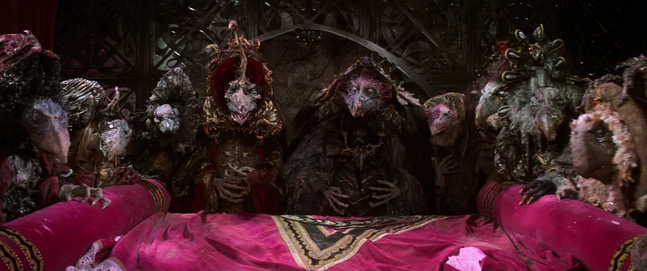 The Dark Crystal (1982) movie still: medium — Seven elaborate bird-like creatures stand behind a long table draped in dark red velvet. They feature…