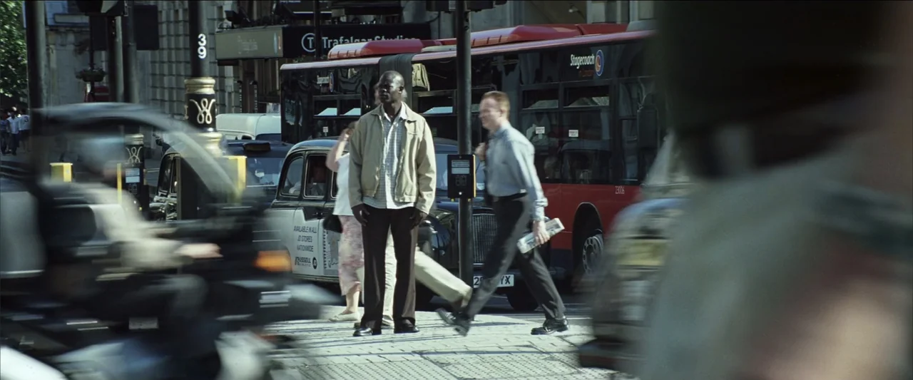 Blood Diamond (2006) movie still: medium — A busy London street corner features a black taxi and a red bus passing. A man stands motionless in the…