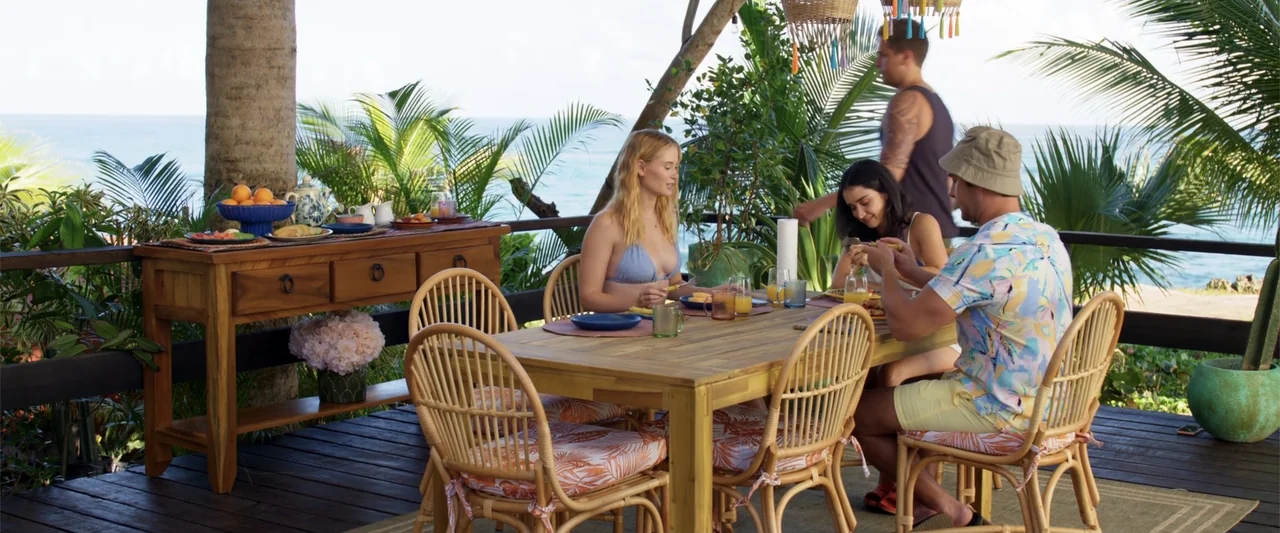 Beautiful Wedding (2024) movie still: medium — An outdoor wooden dining deck features tropical plants overlooking the ocean at daytime. A small…