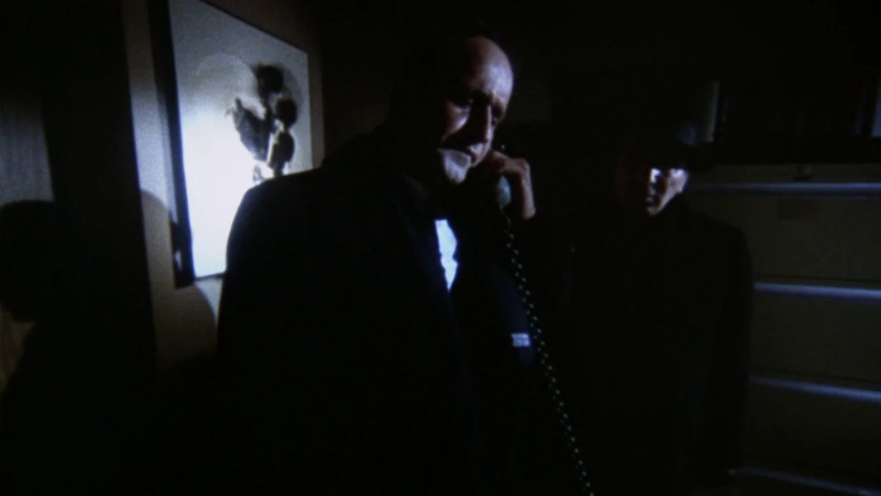 Bullitt (1968) movie still: medium — A dimly lit room contains two men positioned in front of a framed picture. One man holds a coiled telephone…