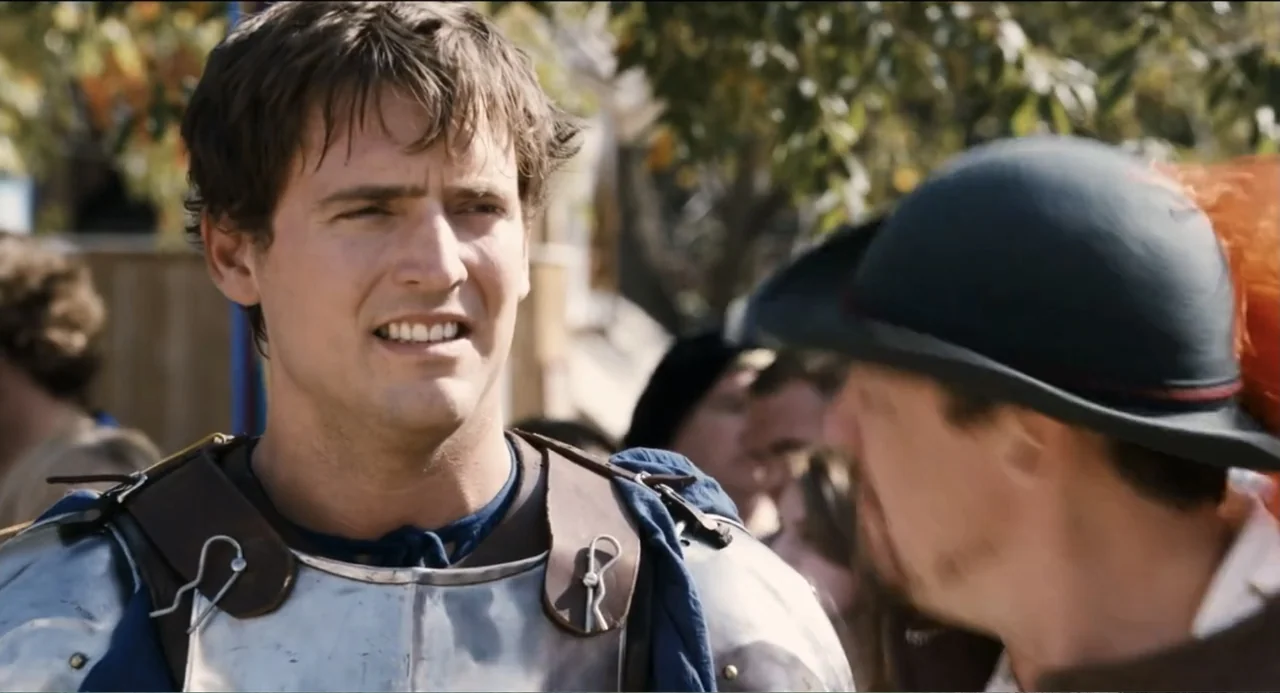 Todo es amor (2024) movie still: medium — An outdoor fairground setting with a crowd of people in the blurred background. A man wearing steel armor…