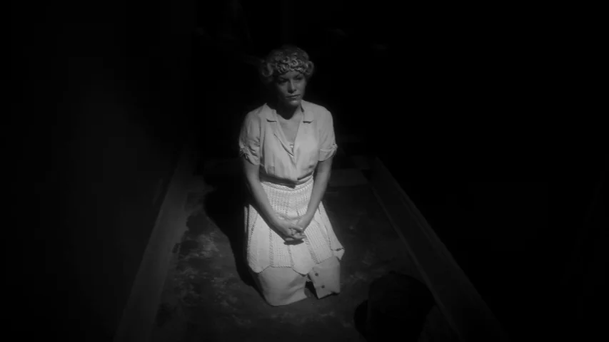 Keyhole (2011) movie still: medium — A dark hallway with a checkered floor is engulfed in heavy shadows. A blonde woman in an apron kneels in a…