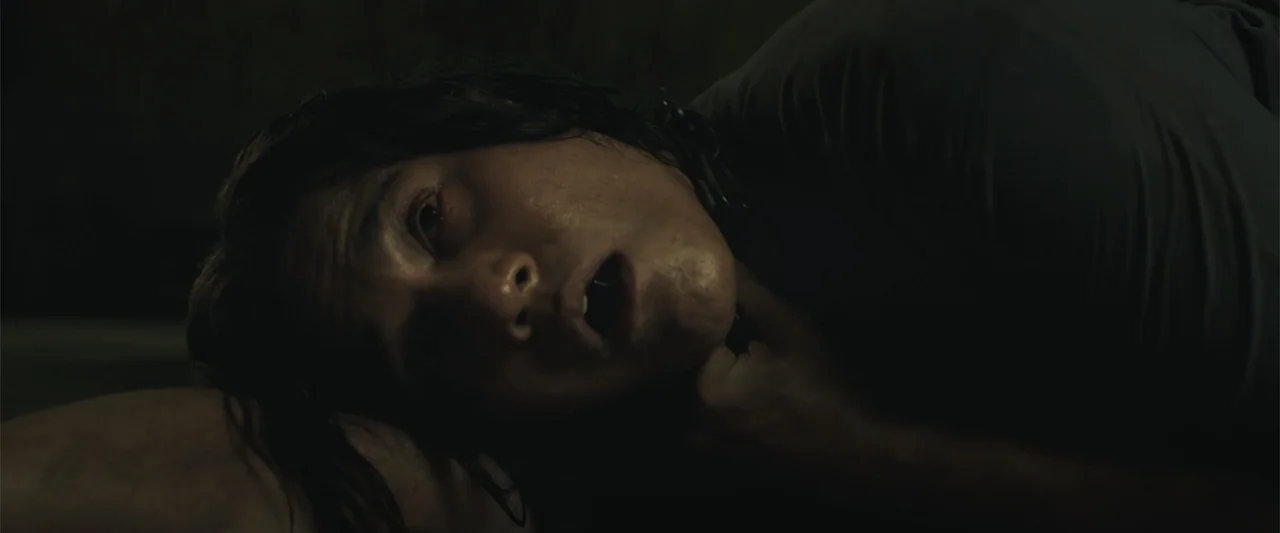 The Unholy (2022) movie still: closeup — A dimly lit room with dark stone-like walls and a rough floor surface. A man lies on the ground in a dark…