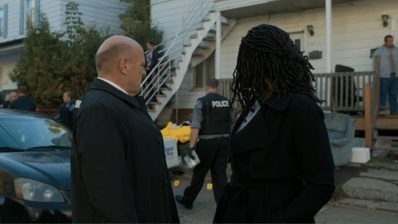 Death Wish (2018) movie still: medium — A residential property exterior with a white house, wooden stairs, and a parked car. Two people stand in the…