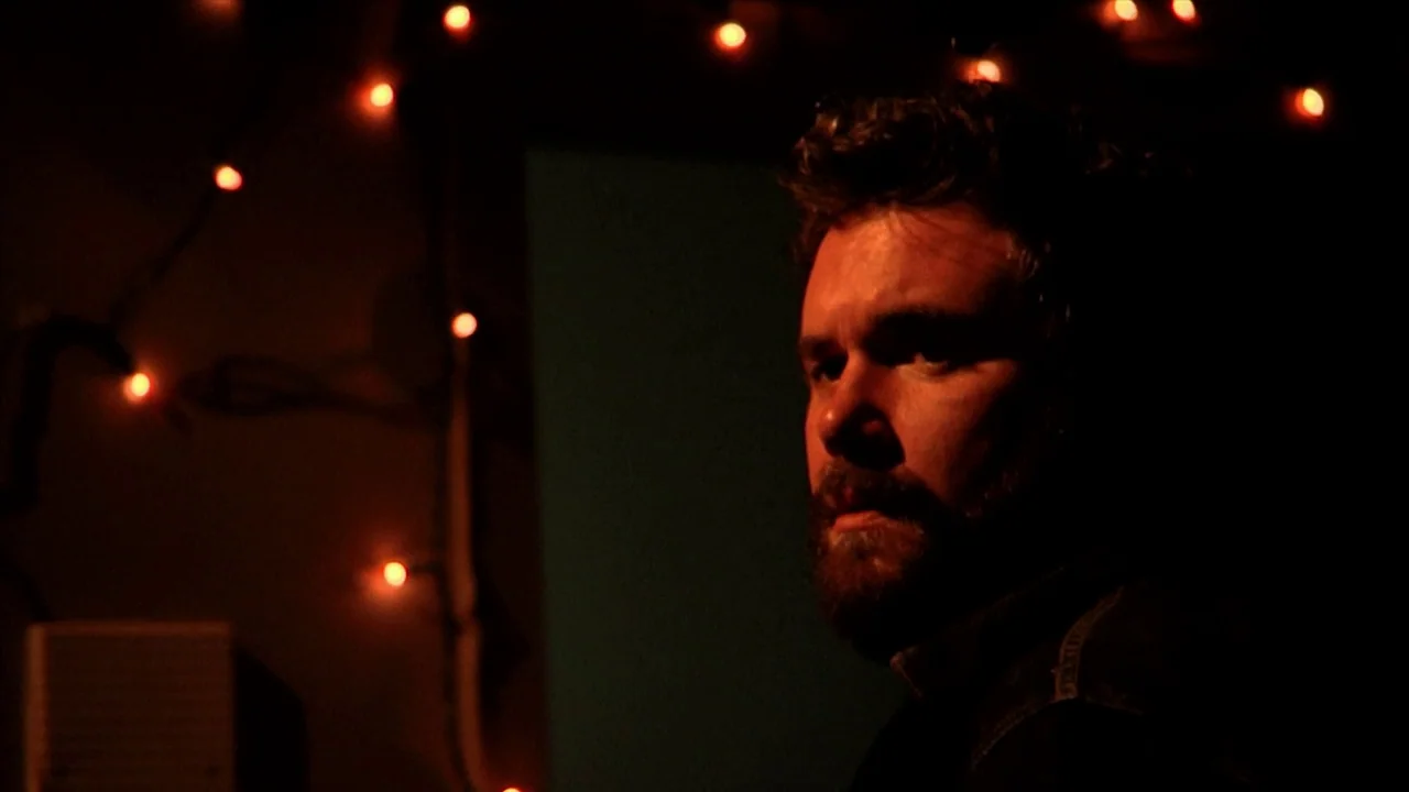 A Horrible Way To Die (2010) movie still: closeup — A dimly lit room features a man partially illuminated by warm light sources. In the background,…