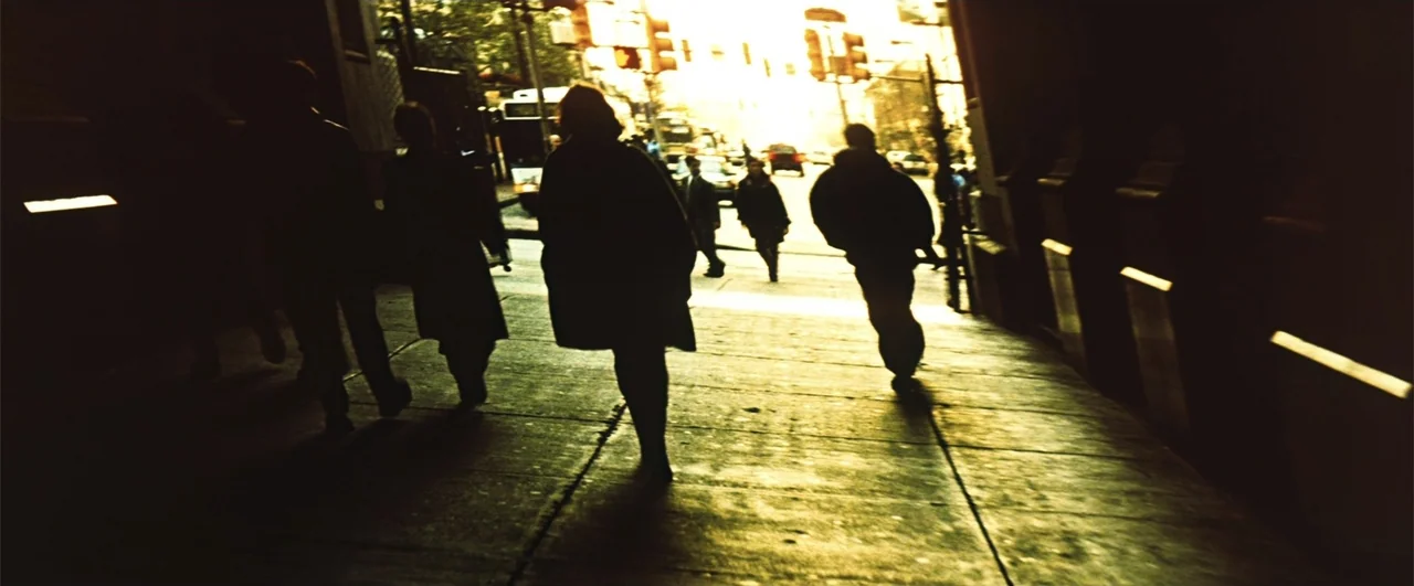 Fallen (2004) movie still: wide — A sidewalk pathway leads into an urban intersection bathed in heavy, bright backlighting. Several individuals in…