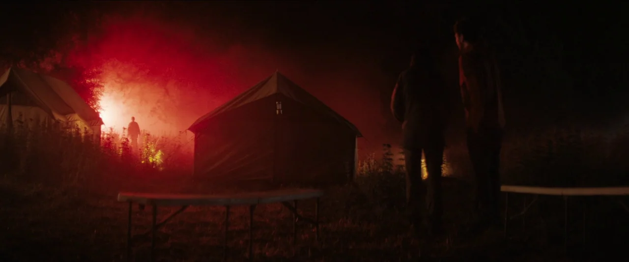 In The Earth (2021) movie still: wide — A dark, nighttime field contains two tents and empty folding tables in the foreground. Two individuals stand…