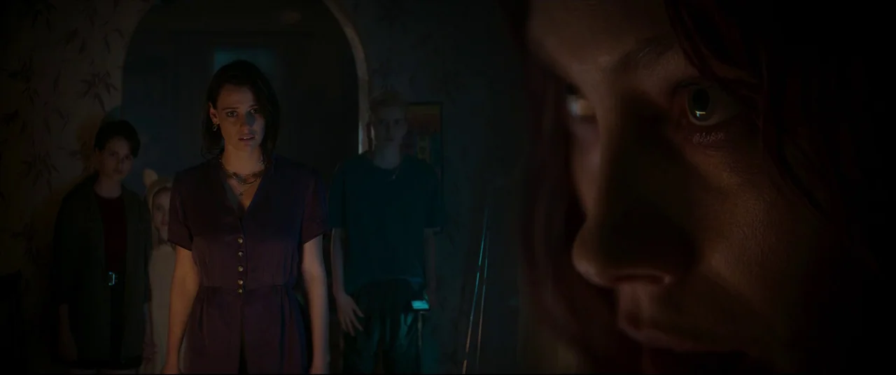 Evil Dead Rise (2023) movie still: medium — A dimly lit hallway features a patterned wallpaper background with an arched doorway. A young woman in a…