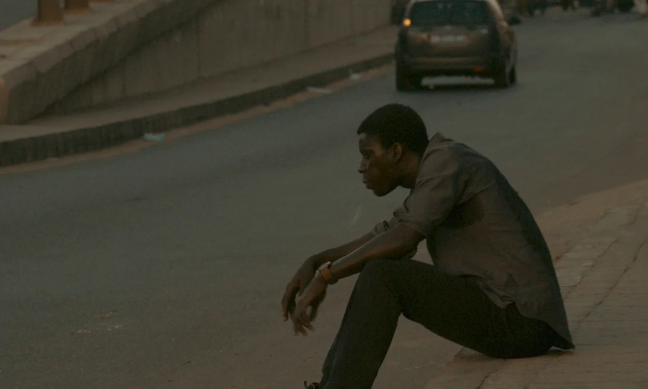 Atlantics (2019) movie still: medium — A paved urban thoroughfare with a concrete curb and a car in the distance. A man sits alone on the curb with…