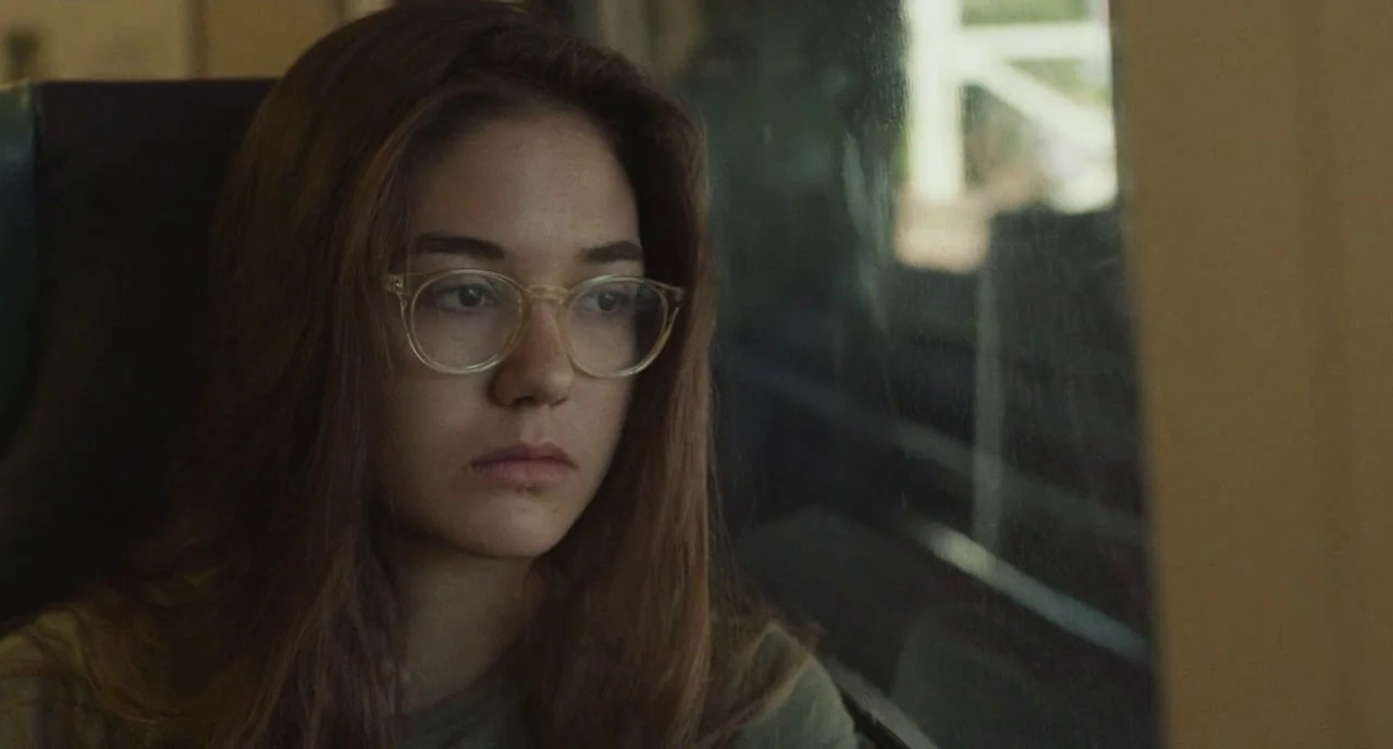 Skate Kitchen (2018) movie still: closeup — A person sits inside a train car next to a large passenger window. The woman gazes outside the glass…