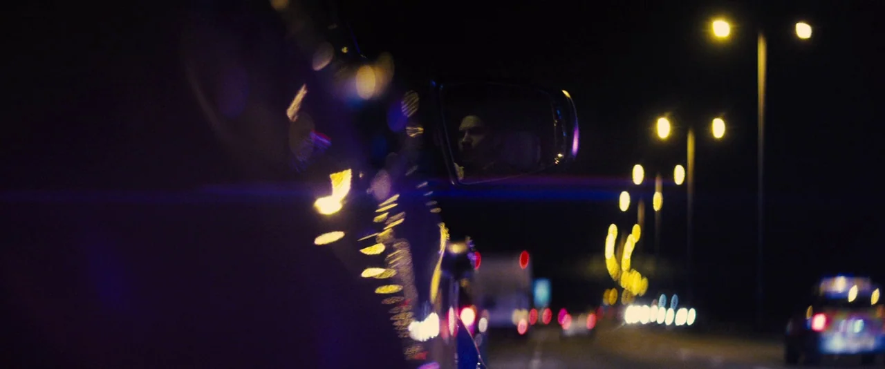 Locke (2013) movie still: closeup — A night view from inside a car driving on a highway with blurred lights. The driver is visible as a reflection…