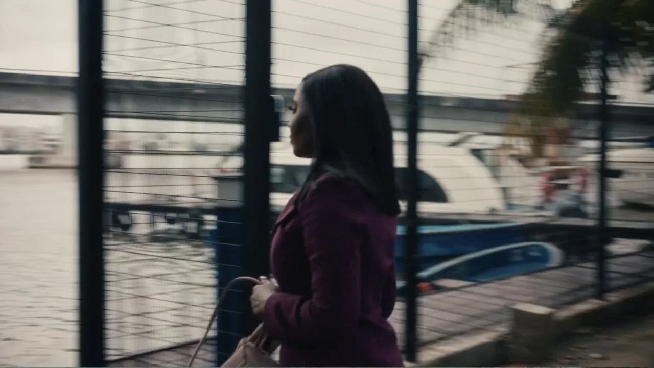 SIN: Blackout (2025) movie still: medium — A woman walks along a metal wire fence next to a boat-filled marina. A large concrete bridge is visible…